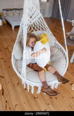 Das kleine Mädchen reitet zu Hause auf dem Makramee-Stuhl auf der Schaukel. Rustikaler Stil im Innenraum. Das Kleinkind trägt Senfhosen und Stiefel aus Cordstoff. Kind ist spielerisch Stockfoto