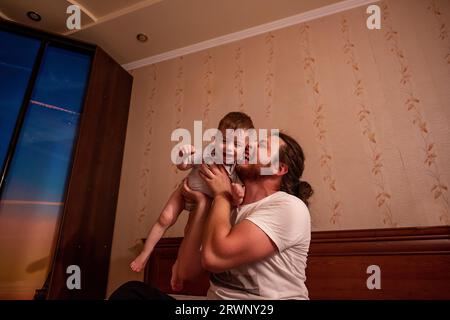 Ein vielseitiger Vater mit langen Haaren und Brille wirft den kleinen Sohn auf das Bett. Ein junger Mann rollt einen Jungen in die Arme, ein Kind gibt vor, ein Flugzeug zu sein Stockfoto