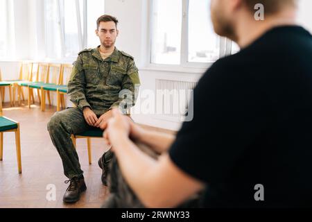 Rückansicht eines strapazierten bärtigen Militärsoldaten in Tarnuniform, der während der Nachkriegsgruppenunterstützungs-Sitzungstherapie im Gespräch ist. Stockfoto