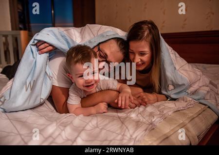 Die fröhliche, süße Familie versteckt sich mit dem kleinen Sohn unter der Decke. Mutter und Vater versuchen, ein aktives Baby zu fangen, der lustige Junge läuft vor den Eltern weg. Junge Ma Stockfoto