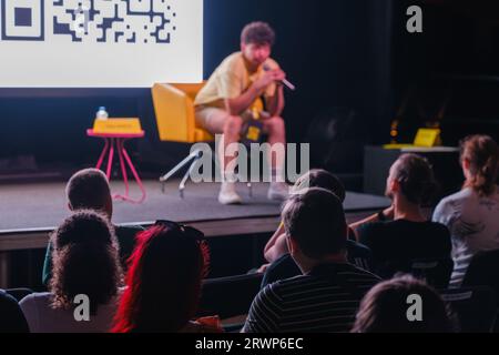 Verschwommener männlicher Trainer, der auf dem Stuhl auf der Bühne sitzt, in der Nähe des Bildschirms und während der Konferenz Fragen der Menge in der dunklen Halle beantwortet Stockfoto