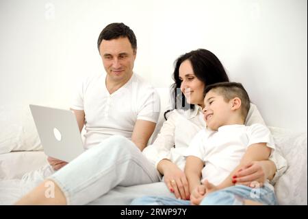 Banner. Neueste Technologie Familie Ehemann Frau und Kind sitzend Film auf Laptop auf dem Bett entspannend weiß cloth.es Chatten online Verwandte Mom Vater und Sohn lachen lächelnd freudig Stockfoto