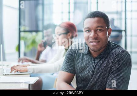 Junger afroamerikanischer Mann in lässiger Kleidung, der vor der Kamera posiert, während er vor dem Hintergrund seiner Kollegen am Tisch sitzt Stockfoto