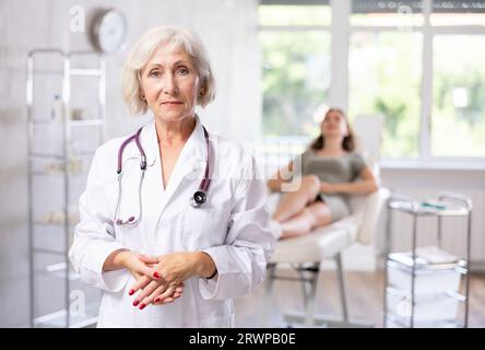 Lächelnde Seniorärztin in Uniform mit dahinter liegender Patientin in der Klinik. Stockfoto