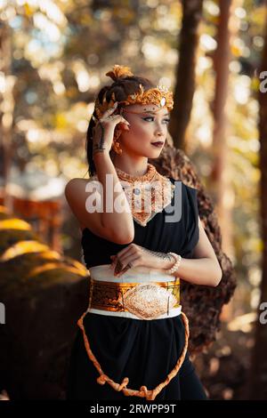 Stammeskönigin im Make-up, während sie eine goldene Krone und eine goldene Halskette mit dem schwarzen Kleid im Wald trägt Stockfoto