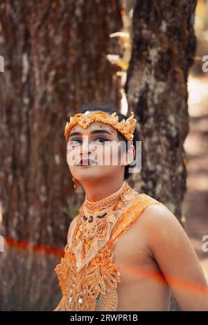 Balinesischer Mann, der im Wald steht, während er bei Tageslicht eine goldene Krone und eine goldene Halskette trägt Stockfoto