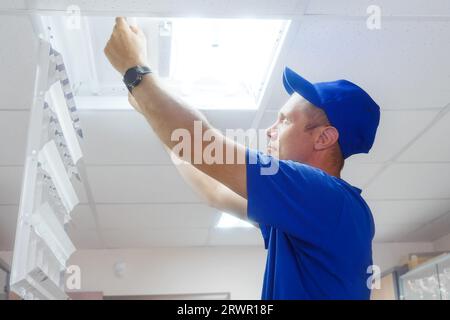 Ein professioneller Elektriker ersetzt die Lampe in der Deckenleuchte. Mann im Overall repariert die Beleuchtung im Büro. Authentischer Workflow. Stockfoto