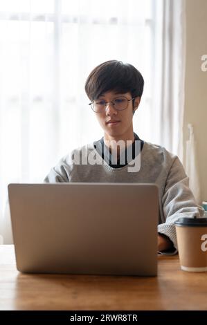 Ein kluger junger asiatischer Student in Brille und Pullover arbeitet an seinem Projekt auf einem Laptop in einem Café. Stockfoto