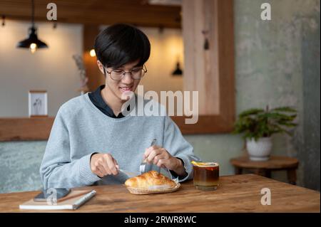 Ein glücklicher junger asiatischer Mann isst gerne ein Croissant in einem wunderschönen Café am Wochenende allein. Lifestyle, Menschen und Lebensmittelkonzept Stockfoto