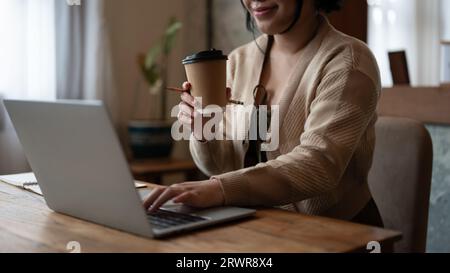 Abgeschnittenes Bild einer schönen und glücklichen jungen asiatischen Frau, die aus der Ferne in einem Café arbeitet und ihren Kaffee genießt, während sie auf ihrem Laptop an einem Tisch arbeitet Stockfoto