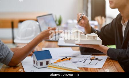 Ein professioneller asiatischer Architekt zeigt einem Kunden ein Hausmodell, während er die Baupläne im Büro diskutiert. Stockfoto