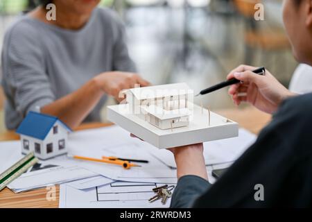 Ein professioneller asiatischer Architekt zeigt einem Kunden ein Hausmodell, während er die Baupläne im Büro diskutiert. Nahaufnahme Stockfoto