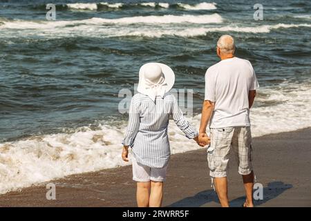 Ein älteres Paar steht am Strand am Meer, schaut auf die Wellen und entspannt sich. Rückansicht von hinten. Stockfoto