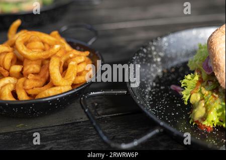 Veganer Burger in geschnittenen und lockigen Pommes frites, serviert in einer dunklen Pfanne, auf einem dunklen Holztisch, Nordrhein-Westfalen, Ruhrgebiet Stockfoto