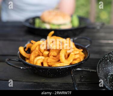 Vegane lockige Pommes frites, serviert in einer dunklen Pfanne, auf einem dunklen Holztisch, im Hintergrund ein Burger und männliche Person, verschwommen, Nordrhein-Westfalen, Ruhr Stockfoto
