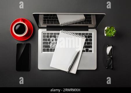 Eleganter schwarzer Büroschreibtisch mit Laptop, Notebook, roter Tasse Kaffee und Mobiltelefon. Draufsicht. Stockfoto