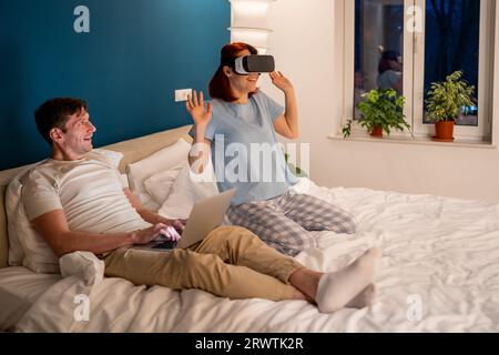 Frau im VR-Headset auf dem Bett, Mann beobachtet, lacht, lächelt. Spaß beim Spielen in der virtuellen Realität Stockfoto