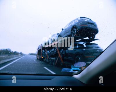 Blick von innen aus einem Auto mit einer mit Wassertropfen bedeckten Windschutzscheibe, wobei der Schwerpunkt auf einem Anhänger mit mehreren Fahrzeugen liegt. Stockfoto