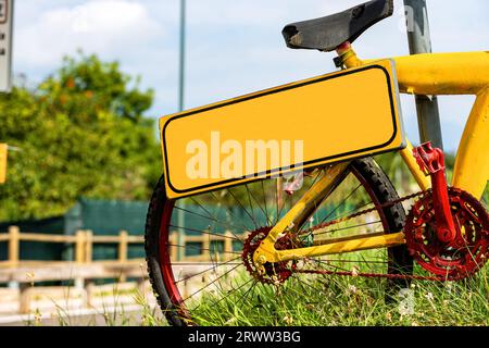 Leeres orangefarbenes Schild mit Kopierraum, der von einem alten Fahrrad hängt, verlassen auf einer Wiese mit grünem Gras. Stockfoto