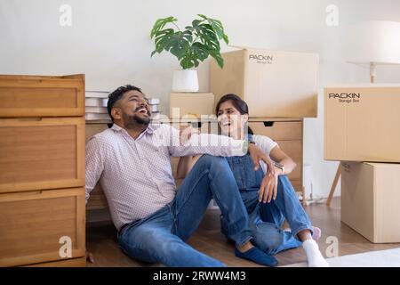 Glückliches junges indisches Paar, das am Umzugstag eine Pause in ein neues Zuhause macht, das auf dem Boden sitzt und von einer Box umgeben ist Stockfoto