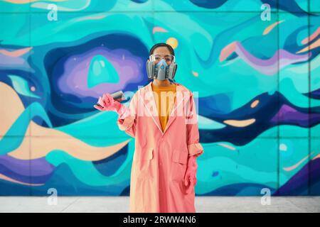 Graffiti-Künstlerin in Atemschutzmaske, die neben der Wand steht, mit ihren Bildern, die auf die Kamera schauen. Street-Art-Konzept Stockfoto