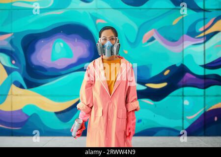 Graffiti-Künstlerin in Atemschutzmaske, die neben der Wand steht, mit ihren Bildern, die auf die Kamera schauen. Street-Art-Konzept Stockfoto