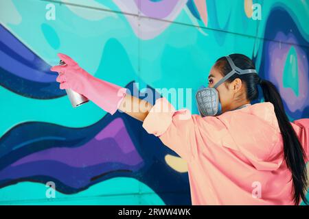 Weibliche Straßenmalerin in Atemmaske Malerei Bunte Graffiti an der Wand Moderne Kunst, städtisches Konzept. Stockfoto