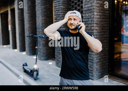 Erwachsener Mann in weißer Baseballmütze und lässiger Kleidung, der in der Nähe eines Elektrorollers steht und auf seinem Mobiltelefon auf der Straße spricht. Der Mann ist verärgert und Stockfoto