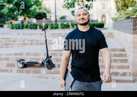 Schöner Mann hält Laptop in der Hand und steht in der Nähe eines Elektrorollers. Aktiver Lebensstil, Rucksacktourist. Ökologie und saubere ökologische Modus der tr Stockfoto