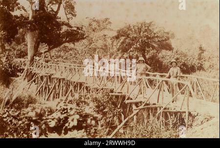 Zwei europäische Männer in Helmen und mehrere afrikanische Männer in kleinen Gruppen stehen auf einer hölzernen Brücke, umgeben von Bäumen und dichtem Laub. Originaltitel: Native Bush Bridge in der Nähe von Accra. Gold Coast 1918. Stockfoto