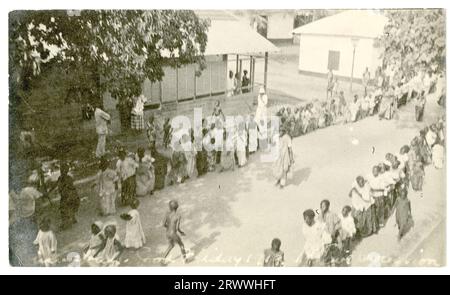 Zwei lange Reihen von Frauen und Kindern ziehen sich durch eine Straße in Winneba, gesäumt von Bäumen und Gebäuden, während andere sehen. Alle sind elegant gekleidet. Eine kleine Gruppe von Kindern in weißen Kleidern und Hauben läuft zwischen den Linien die Mitte der Straße hinauf. Im Negativen: Winnebah. Karfreitag 1919. Einheimische Prozession. Stockfoto