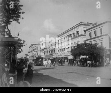 Straßenszene gesäumt von Kolonialgebäuden. Europäische und indische Fußgänger laufen oder reisen in Pferdekutschen. Stockfoto