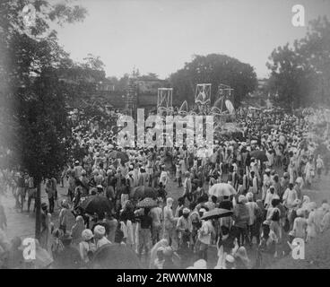 Religiöse Prozession mit geschmückten Wagen inmitten eines großen Festes von traditionell gekleideten Indianern. Bäume säumen den Platz und Gebäude sind im Hintergrund zu sehen. Stockfoto