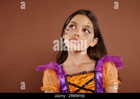 Nachdenkliches Mädchen in farbenfrohem Kostüm mit Halloween-Make-up auf braunem Hintergrund, Oktober Stockfoto