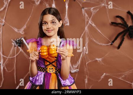 Fröhliches Mädchen mit Spinnennetz-Make-up im Halloween-Kostüm mit Kürbissen auf braunem Hintergrund Stockfoto