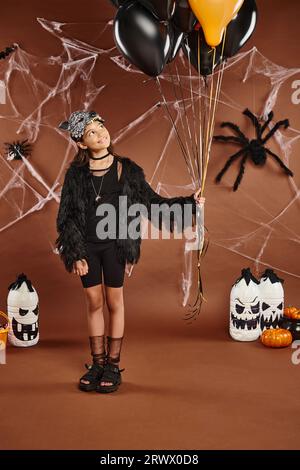 Smiley Girl hält schwarze und orangefarbene Ballons auf braunem Hintergrund mit Netz und Spinnen, Halloween Stockfoto