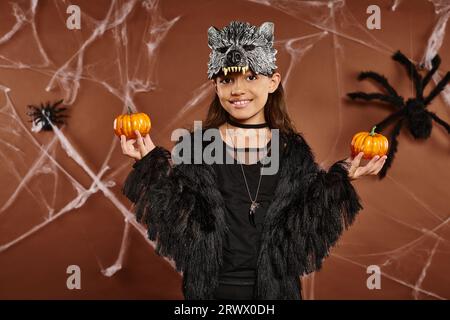 Smiley Girl in Wolf Maske hält Kürbisse auf braunem Hintergrund mit Spinnen und Spinnennetz, Halloween Konzept Stockfoto