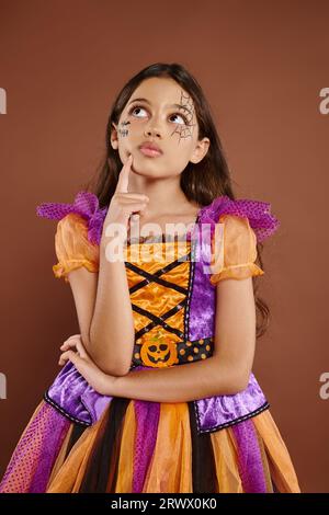 Nachdenkliches Mädchen in farbenfrohem Kostüm mit Halloween-Make-up auf braunem Hintergrund, Oktober Stockfoto