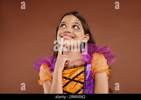 Traumhaftes Mädchen in farbenfrohem Kostüm mit Halloween-Make-up, das auf braunem Hintergrund wegschaut, fröhliches Gesicht Stockfoto