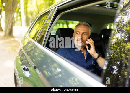 Afrikanischer Geschäftsmann, der Smartphone hält, während er die Kamera anschaut. Erfolgreicher Unternehmer, der blaue Krawatte trägt und im Auto mit dem Mobiltelefon sitzt. Hap Stockfoto