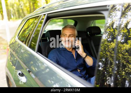 Afrikanischer Geschäftsmann, der Smartphone hält, während er die Kamera anschaut. Erfolgreicher Unternehmer, der blaue Krawatte trägt und im Auto mit dem Mobiltelefon sitzt. Hap Stockfoto