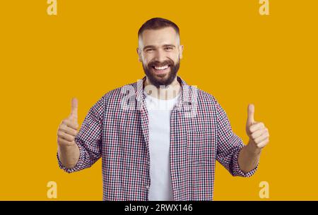 Junger brünettbärtiger Mann in kariertem Hemd mit Daumen auf gelbem Hintergrund und Blick auf die Kamera. Stockfoto