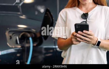 Frau, die ihr Handy benutzt, um ihr Elektroauto aufzuladen. EV-Station und mobiles Bezahlkonzept. Stockfoto