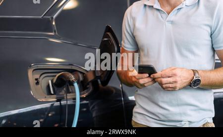 Männliche Person, die sein Smartphone zum Laden eines Elektroautos verwendet. EV und mobiles Bezahlkonzept. Stockfoto