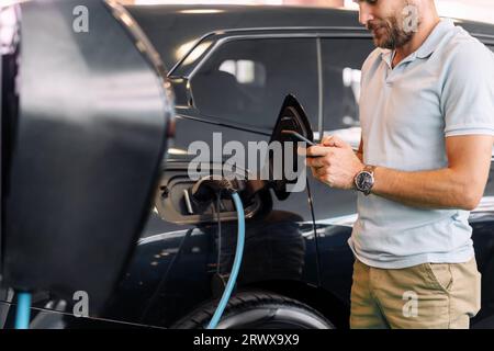 Junger Erwachsener, der für das Aufladen des Elektrofahrzeugs auf dem Mobiltelefon bezahlt. Geschäftsmann, der Freizeitkleidung trägt und sein Handy vor einem schwarzen Elektroauto benutzt. Stockfoto