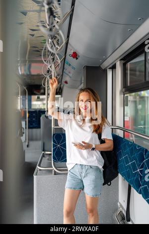 Attraktive junge Erwachsene, brünette Frau steht im Trolley-Bus und schaut auf die Kamera und lächelt. Weibliche Person mit persönlichem Informationsgerät Stockfoto