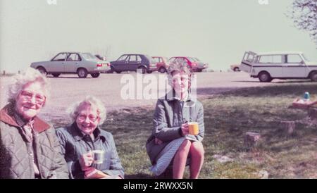 Archivfoto von drei älteren Damen an einem Picknickplatz bei einer Tasse Tee aus einer Flasche mit alten Autos auf dem Parkplatz im Hintergrund, Ende der 1970er Jahre Stockfoto
