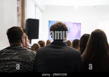 Hintergrund nicht erkennbarer Personen, die mit dem Rücken zur Kamera stehen und der Bühne zugewandt sind, auf der die Lautsprecher sprechen. Geschäftsereignis. Hochwertige Fotos Stockfoto