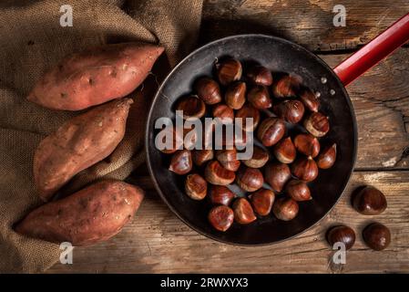 Blick von oben auf einige Kastanien in einer Pfanne, die zum Braten vorbereitet ist, und einige Süßkartoffeln auf einem alten Holztisch Stockfoto