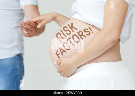 Eine schwangere Frau hält die Hand ihres Mannes auf dem Bauch mit der Inschrift - Risikofaktoren, ein Ausrufezeichen. Schwangerschaftskonzept. Stockfoto